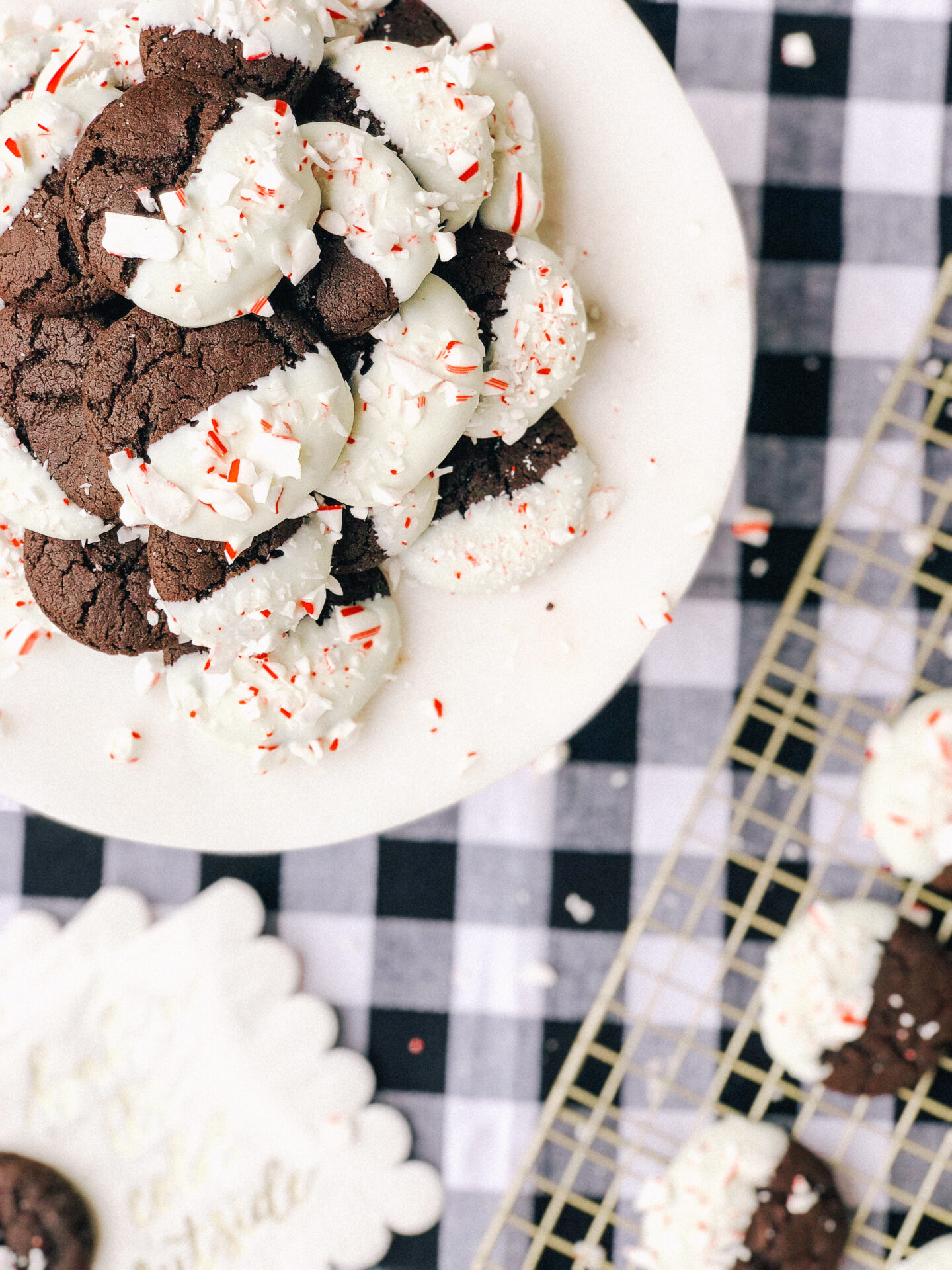 dark chocolate cookies dipped in white chocolate and topped with crushed peppermint on gold cooling rack and white cake stand on black and white check tablecloth
